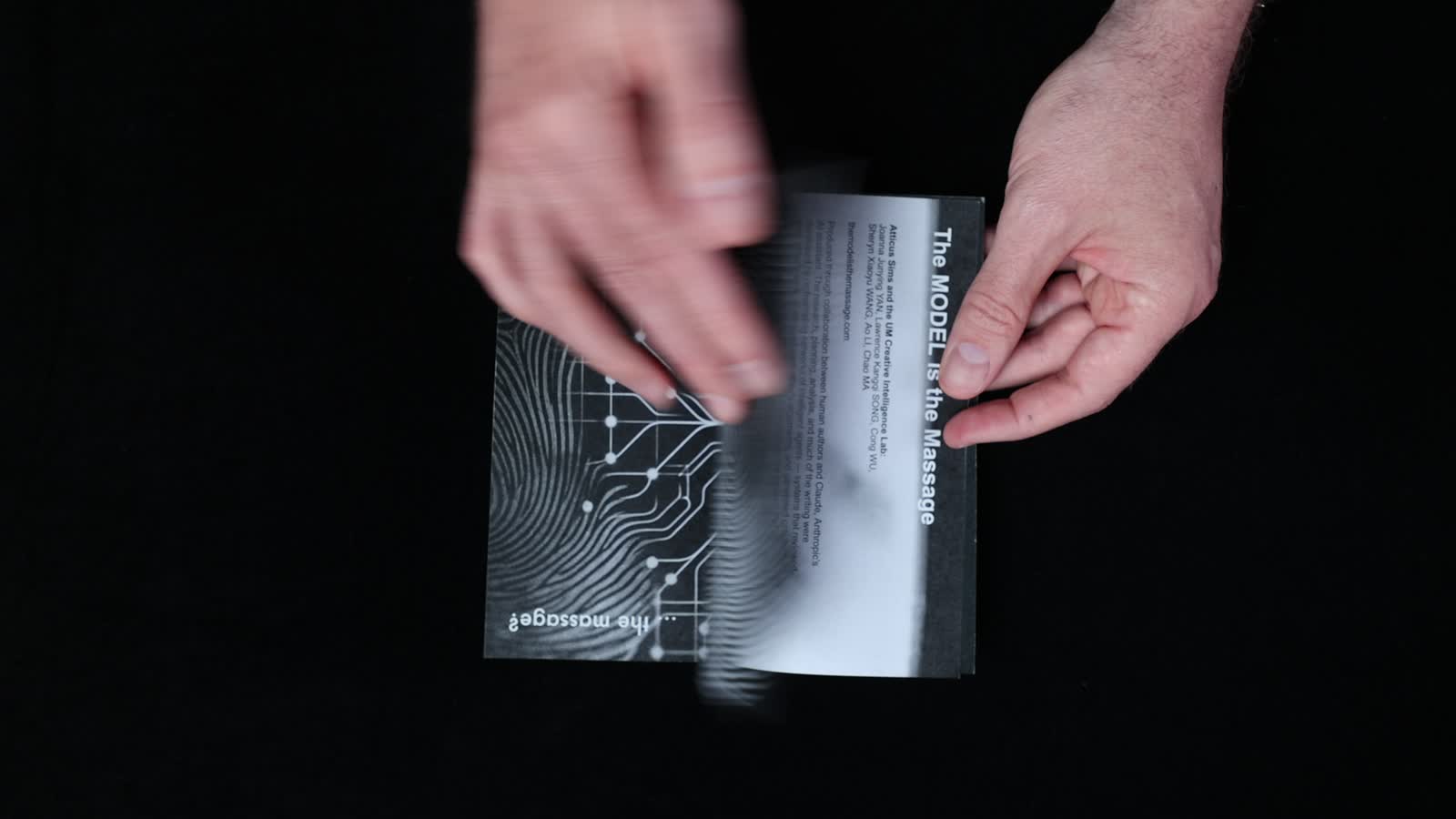 Hands turning pages of the actual printed book on a dark surface.